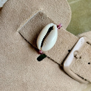 May include: A beige suede sandal strap featuring a white cowrie shell centred on a thin string of small, translucent pink beads. The strap includes a matching beige buckle and a rectangular cutout, showcasing a textured, soft material finish.
