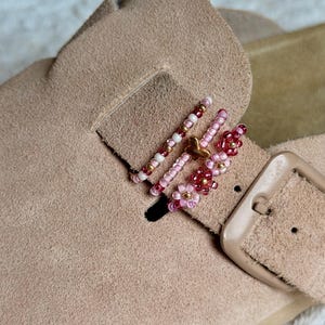 Könnte beinhalten: Ein beigefarbener Slipper aus Wildleder mit einem dekorativen Riemen, der mit drei Perlenringen verziert ist. Die Ringe bestehen aus einer Mischung aus rosa, weißen und goldfarbenen Perlen, ergänzt durch einen kleinen Schmetterling und Blumen.