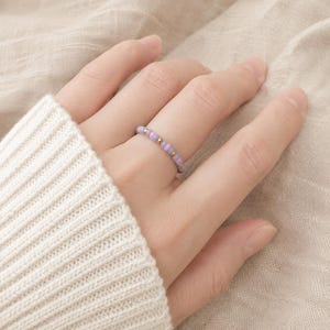 Könnte beinhalten: Ein zarter Ring mit einem sich wiederholenden Muster aus kleinen, runden, hellvioletten und lavendelfarbenen Perlen, die mit winzigen, polierten goldfarbenen Metallperlen durchsetzt sind und ein schlichtes, farbenfrohes Ringdesign bilden.