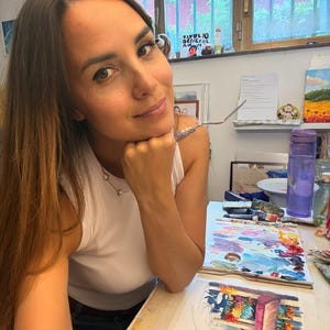 May include: A white sleeveless top worn with dark denim shorts. On the desk, there is a palette with various oil paint colors, a painting of a flower-filled storefront, a purple water bottle, and a metal palette knife held near the chin.