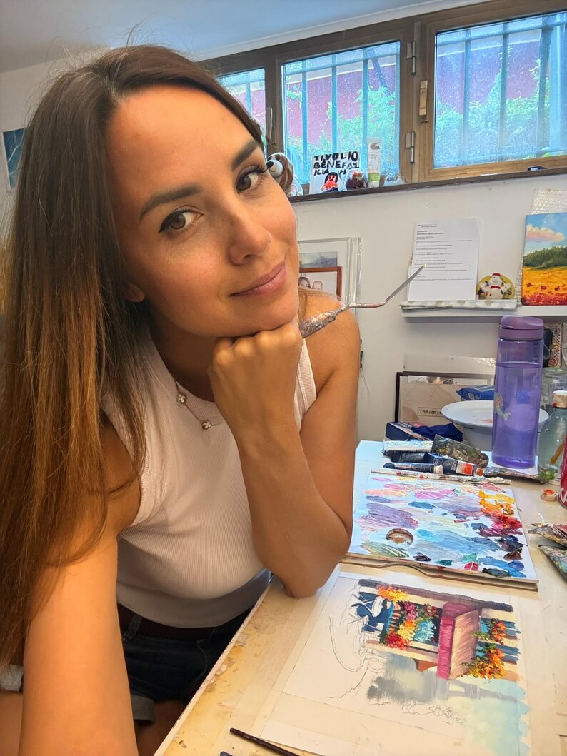 May include: A white sleeveless top worn with dark denim shorts. On the desk, there is a palette with various oil paint colors, a painting of a flower-filled storefront, a purple water bottle, and a metal palette knife held near the chin.