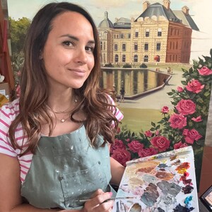 May include: An oil painting on canvas depicting a large stone building with a dark roof situated behind a reflective pond, framed by vibrant pink rose bushes in the foreground, held by an artist wearing a sage green apron and striped shirt.