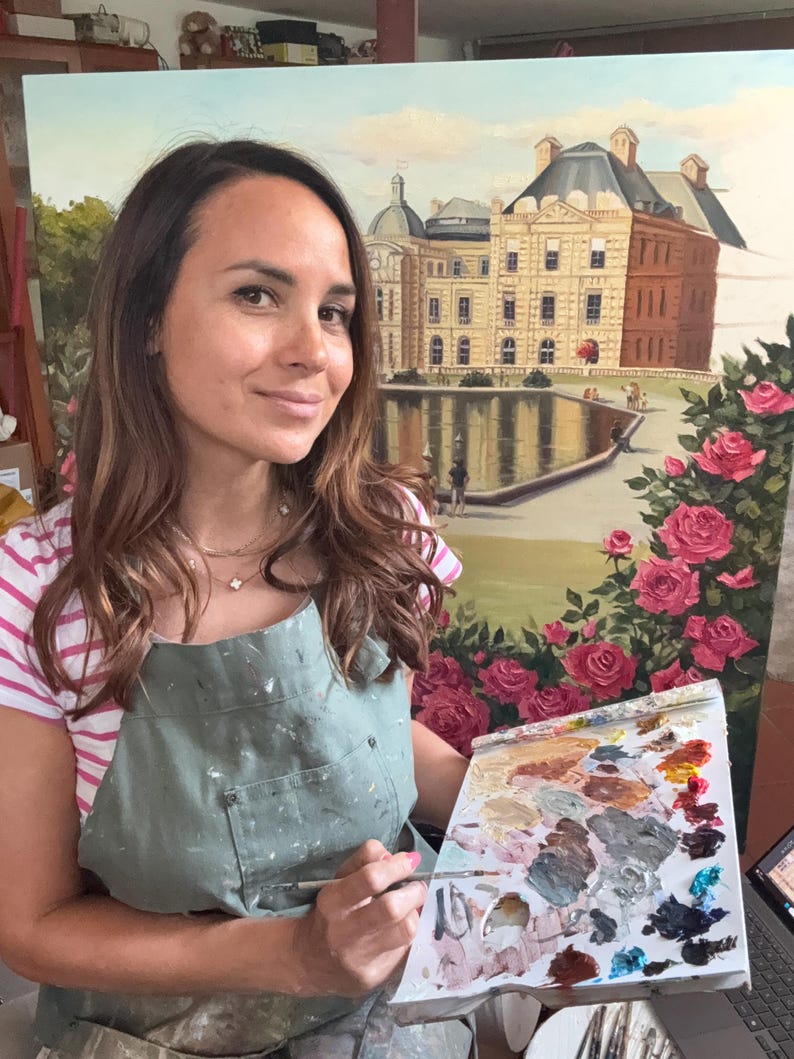 May include: An oil painting on canvas depicting a large stone building with a dark roof situated behind a reflective pond, framed by vibrant pink rose bushes in the foreground, held by an artist wearing a sage green apron and striped shirt.