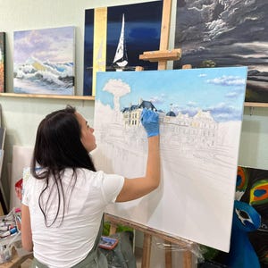 May include: A large rectangular canvas on a wooden easel features an oil painting in progress, depicting a detailed architectural scene with a fountain and buildings under a blue sky with soft white clouds, painted with blue and white tones.