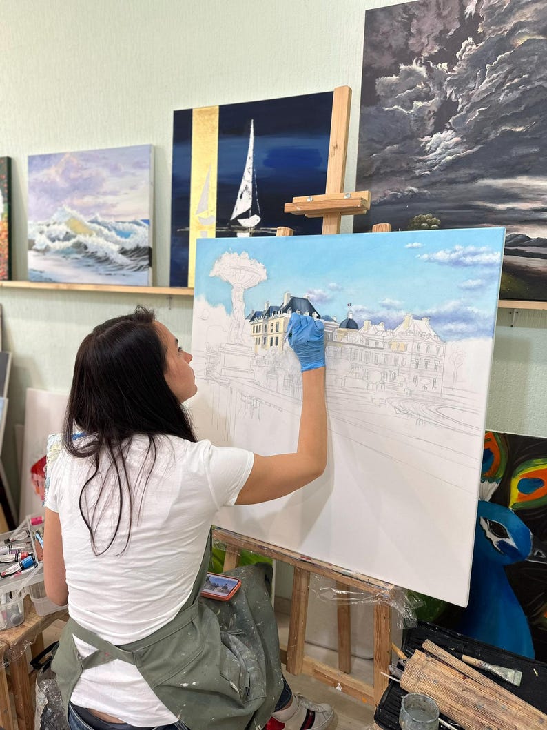 May include: A large rectangular canvas on a wooden easel features an oil painting in progress, depicting a detailed architectural scene with a fountain and buildings under a blue sky with soft white clouds, painted with blue and white tones.
