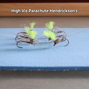 May include: Two fishing flies with bright chartreuse parachute wings and brown bodies. The hooks are curved and silver. The text "High Vis Parachute Hendrickson's" is above the flies.