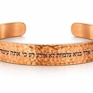 May include: A copper-colored metal cuff bracelet with a hammered, textured finish. The exterior surface features black engraved Hebrew text that reads "גם כי אלך בגיא צלמות לא אירא רע כי אתה עמדי" flanked by two small arrow symbols on either side of the inscription.