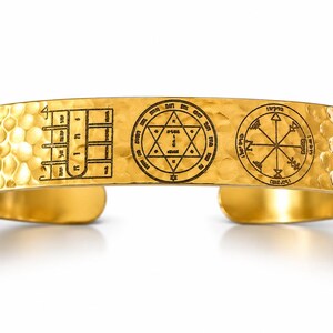 Puede incluir: Un brazalete rígido de tono dorado con textura martillada. La superficie presenta tres símbolos negros grabados: una cuadrícula rectangular con caracteres hebreos, una estrella de seis puntas dentro de un círculo y un emblema circular.