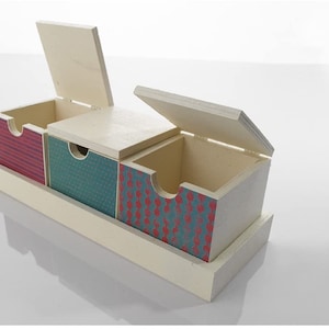May include: A wooden storage box with three compartments. Each compartment has a different patterned design. The box has a cream-coloured base and lids, with red and teal patterns on the compartments. The lids are open, revealing the interior.