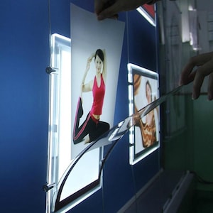 May include: Illuminated display panels mounted on a blue wall, each showcasing a photograph. One panel displays a woman in a yoga pose. The panels are backlit, creating a bright, modern aesthetic. A photograph is being inserted into the panel.