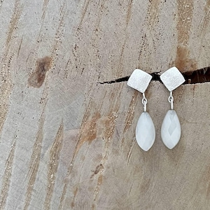May include: A pair of silver earrings with white teardrop-shaped stones. The earrings have a square-shaped design at the top.