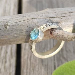 May include: A silver ring with a light blue gemstone set in the center. The ring has a textured band and is set on a wooden surface.