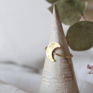 Può includere: Un anello a forma di mezzaluna dorata esposto su un porta anelli in legno bianco. L'anello è semplice e delicato, con una texture martellata.