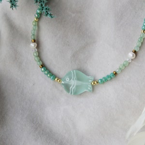 Può includere: Una collana delicata con un ciondolo a forma di pesce verde chiaro, con un contorno bianco. La collana è realizzata con perline verdi e bianche, tra cui una perla.