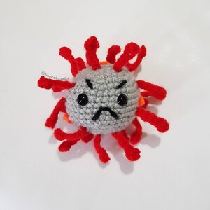 Pode incluir: Um brinquedo de pelúcia de crochê em forma de vírus com um exterior vermelho e laranja espinhoso e um rosto cinza com uma expressão de carranca. O brinquedo tem olhos pretos e uma boca preta. O texto "@hooked.crafts" é visível no fundo.