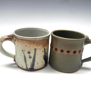 May include: Two ceramic mugs, one with a white glaze and brown and black splatter paint design, and the other with a green glaze and a brown band with small brown dots.