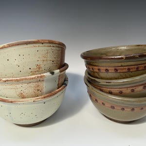 May include: Two stacks of handmade ceramic bowls. One stack features bowls with a speckled cream and brown glaze. The other stack has bowls with a green and brown glaze, accented with a brown band and dotted design.