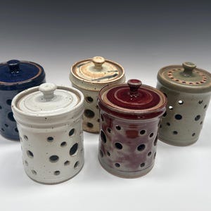 May include: Five ceramic garlic keepers in various colors: navy blue, cream, tan, burgundy, and olive green. Each has a matching lid and is perforated with circular holes for ventilation. The keepers are arranged on a white surface.