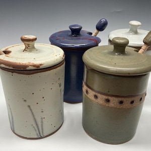 May include: Four ceramic honey pots with lids and wooden dippers. The pots are in various colors, including white, blue, green, and brown. The dippers are all natural wood.