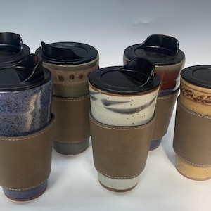 May include: A collection of ceramic travel mugs with black lids and brown leather sleeves. The mugs have various colors and patterns, including blue, brown, and speckled designs. The sleeves are stitched with white thread.