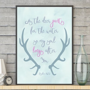 Könnte beinhalten: Ein gerahmter Druck mit hellblauem Aquarellhintergrund zeigt den Text "As the deer pants for the water so my soul longs after You" in Schriftform. Graue Geweihe und der Text "Psalm 42,1" sind ebenfalls enthalten. Dekorative Flaschen und eine Vase stehen auf einem Regal.