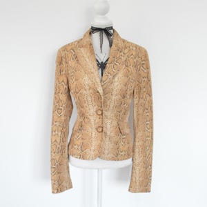 May include: A tan and brown blazer featuring a snakeskin pattern throughout. The garment has long sleeves, a notched lapel collar, and two front buttons for closure. A black lace ribbon is tied in a bow around the neck of the mannequin.