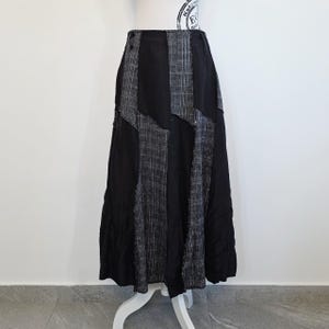 Può includere: Una gonna lunga nera con pannelli verticali in tessuto a quadri grigio e bianco. La gonna è esposta su un manichino bianco, mostrando il suo design fluido e asimmetrico. La gonna ha una vita alta e una silhouette svasata.