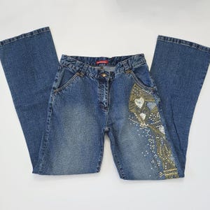 Puede incluir: Vaqueros azules con pernera acampanada. Los vaqueros presentan bordados decorativos con diseños de corazón y flores en oro, plata y blanco. Los vaqueros están colocados sobre una superficie blanca.