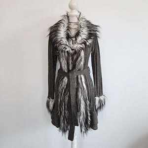 Könnte beinhalten: Langer, dunkelgrauer Cardigan mit Kunstfellkragen und -bündchen. Der Cardigan hat einen passenden Gürtel und weiße und schwarze Kunstfellakzente auf der Vorderseite. Das Kleidungsstück wird auf einer weißen Schaufensterpuppe präsentiert.