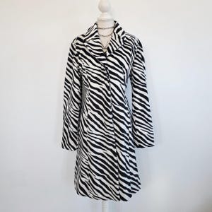 May include: A long-sleeved coat featuring a black and white zebra stripe pattern. The coat has a notched collar, a button-front closure, and a tailored silhouette that extends to the mid-thigh, displayed on a white mannequin torso.