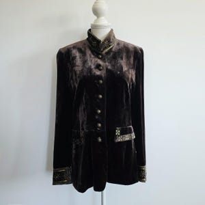 Puede incluir: Chaqueta de terciopelo marrón oscuro con cuello alto y mangas largas. La chaqueta presenta botones dorados decorativos y adornos en el cuello, los bolsillos y los puños. La chaqueta se exhibe en un maniquí blanco.