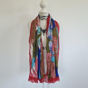 May include: A colorful scarf with a floral pattern in shades of red, blue, green, and pink. The scarf is draped over a white mannequin. The scarf has a red border and a pleated texture.