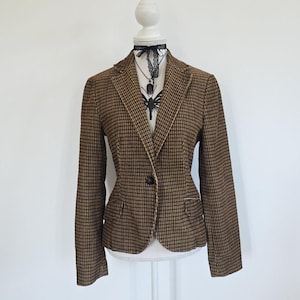 Pode incluir: Um blazer xadrez castanho e preto com fecho de um botão, lapelas entalhadas e dois bolsos frontais com aba. O blazer é exibido num manequim branco, que também usa um colar tipo gargantilha de renda preta com um pendente escuro.