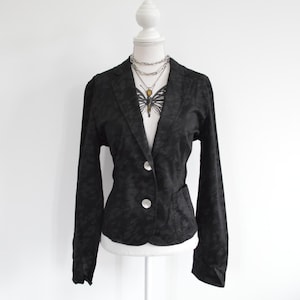 May include: A black blazer featuring a subtle tonal animal print pattern, long sleeves, a notched lapel, and two large, round, iridescent buttons. The garment is displayed on a white mannequin, layered over a white top with a butterfly graphic.