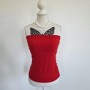 Op de afbeelding: Een felrode strapless tube-top met een zwarte vlinderprint op de borst. Het kledingstuk wordt getoond op een etalagepop en is voorzien van een witte kralenketting die rond de nek van de etalagepop rust.