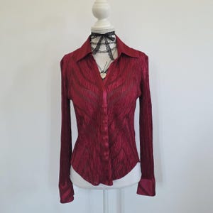 Op de afbeelding: Een bordeauxrode blouse met lange mouwen en een textuur met plooien. De blouse heeft een kraag en een V-hals. De manchetten en kraag zijn donkerder bordeauxrood. Een zwarte kanten choker en ketting sieren de hals van de paspop.