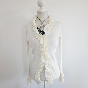Op de afbeelding: Crèmekleurig shirt met lange mouwen en ruches aan de voorkant en rond de kraag. Een zwarte ketting met een zilveren hanger en een vlinderhanger wordt op de shirt getoond. Het shirt staat op een witte mannequin.