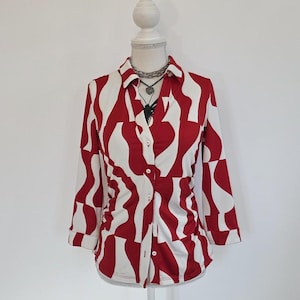Puede incluir: Una camisa de manga larga con botones que presenta un patrón abstracto audaz de formas orgánicas rojas y blancas. La camisa tiene un cuello, una tapeta frontal con botones blancos y mangas de tres cuartos de largo con puños.