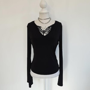 Könnte beinhalten: Ein schwarzes Langarmshirt mit V-Ausschnitt und einer silberfarbenen Kette. Der Ausschnitt verfügt über einen Ausschnitt, der einen schwarz-weißen Schmetterlingsgrafikdruck freigibt. Das Kleidungsstück wird auf einer weißen Schaufensterpuppe präsentiert.