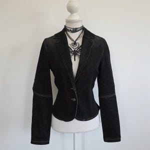 May include: A dark denim blazer with a single button fastening. The jacket has a notched collar, long sleeves, and visible stitching. A decorative necklace with a butterfly pendant is visible.