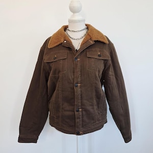 May include: A brown corduroy jacket with a tan sherpa collar. The jacket features two chest pockets, button fastenings, and long sleeves. The jacket is displayed on a mannequin.