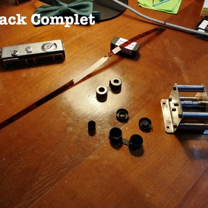 Peut inclure: Un rembobineur de film argenté avec un bouton noir, un rouleau de film et un appareil photo argenté avec un étui noir. Le rembobineur de film est sur une surface en bois avec des boîtes de film noires et un rouleau de film.