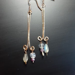 May include: A pair of handcrafted earrings with gold-colored wire and small, colorful gemstones. The earrings feature a long, slender design with a hook closure. Each earring has a unique combination of clear, purple, and iridescent stones.