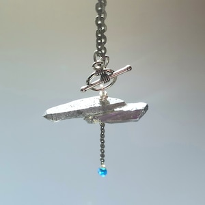 May include: A silver chain necklace with a silver toggle clasp and a raw crystal pendant. A small blue gemstone dangles from the bottom of the pendant.