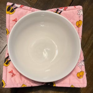 May include: A white ceramic bowl rests on a pink bowl cosy. The cosy features a pattern of cartoon characters in yellow, black, red, and white. The cosy is square with a quilted design.