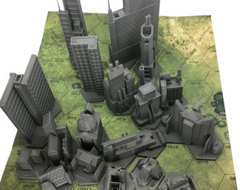May include: A set of 15 grey 3D printed miniature buildings, including skyscrapers, towers, and other structures. The buildings are arranged on a green hexagonal game board with numbers printed on each hexagon.