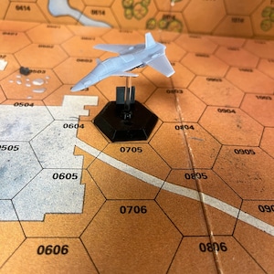 May include: A white plastic model airplane on a black base, sitting on a brown hexagonal game board with white numbers.