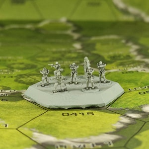 May include: A set of seven white plastic miniature figures, each holding a weapon, standing on a grey circular base. The figures are arranged in a circle on the base, with the base sitting on a green and brown surface.
