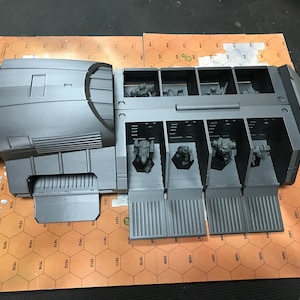 Pode incluir: Modelo cinza, impresso em 3D, de um veículo de transporte com vários compartimentos, cada um abrigando figuras em miniatura. O modelo está sobre um mapa de grade hexagonal, sugerindo um jogo de tabuleiro. O veículo tem um design detalhado.