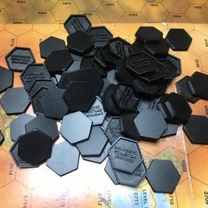 Op de afbeelding: Een stapel zwarte zeshoekige spelstukken met tekst op sommige van hen. De stukken liggen op een bruin en gouden spelbord met zeshoekige rasterquadraten.
